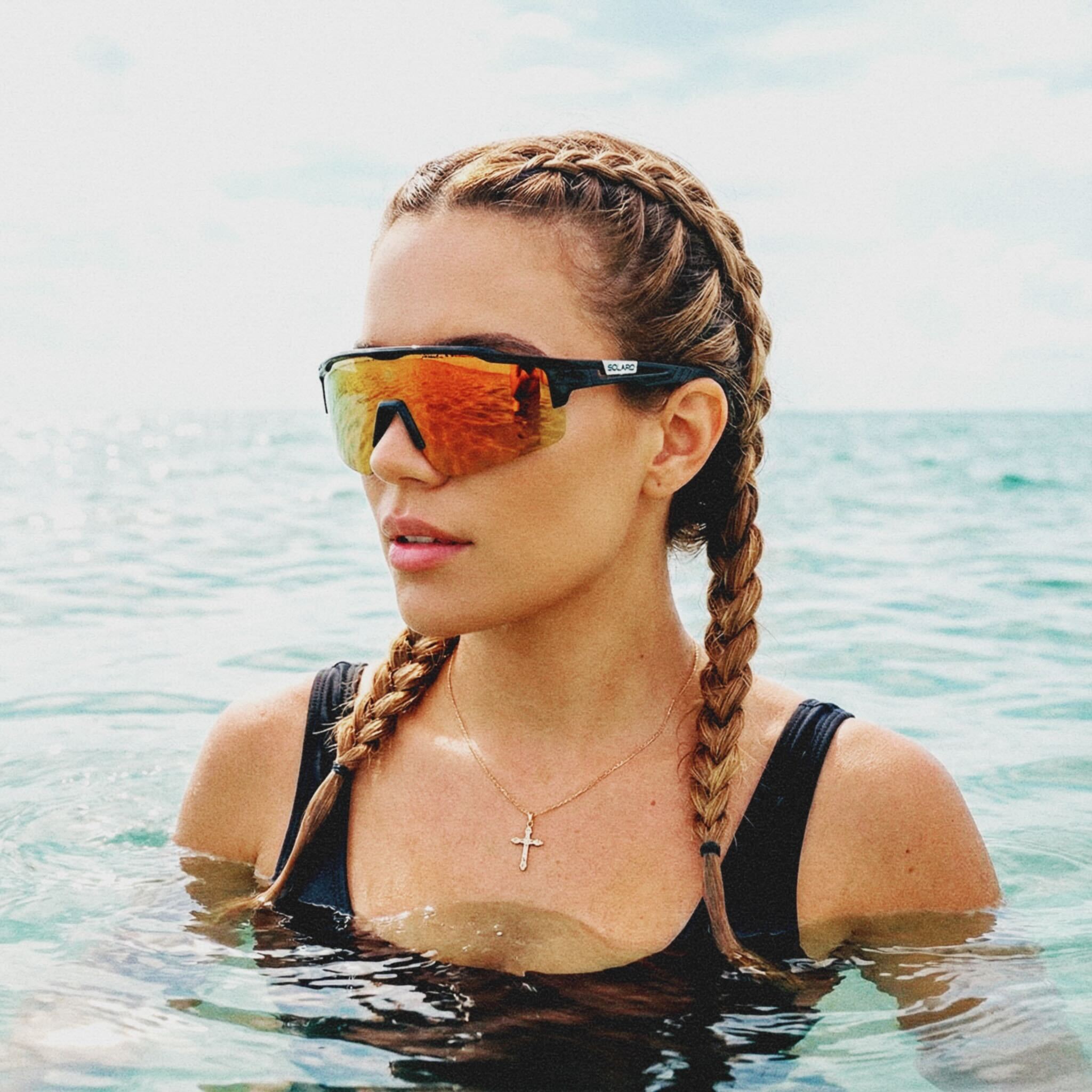 Woman with braided hair wearing Solaro shades sunglasses and a black swimsuit in the water.