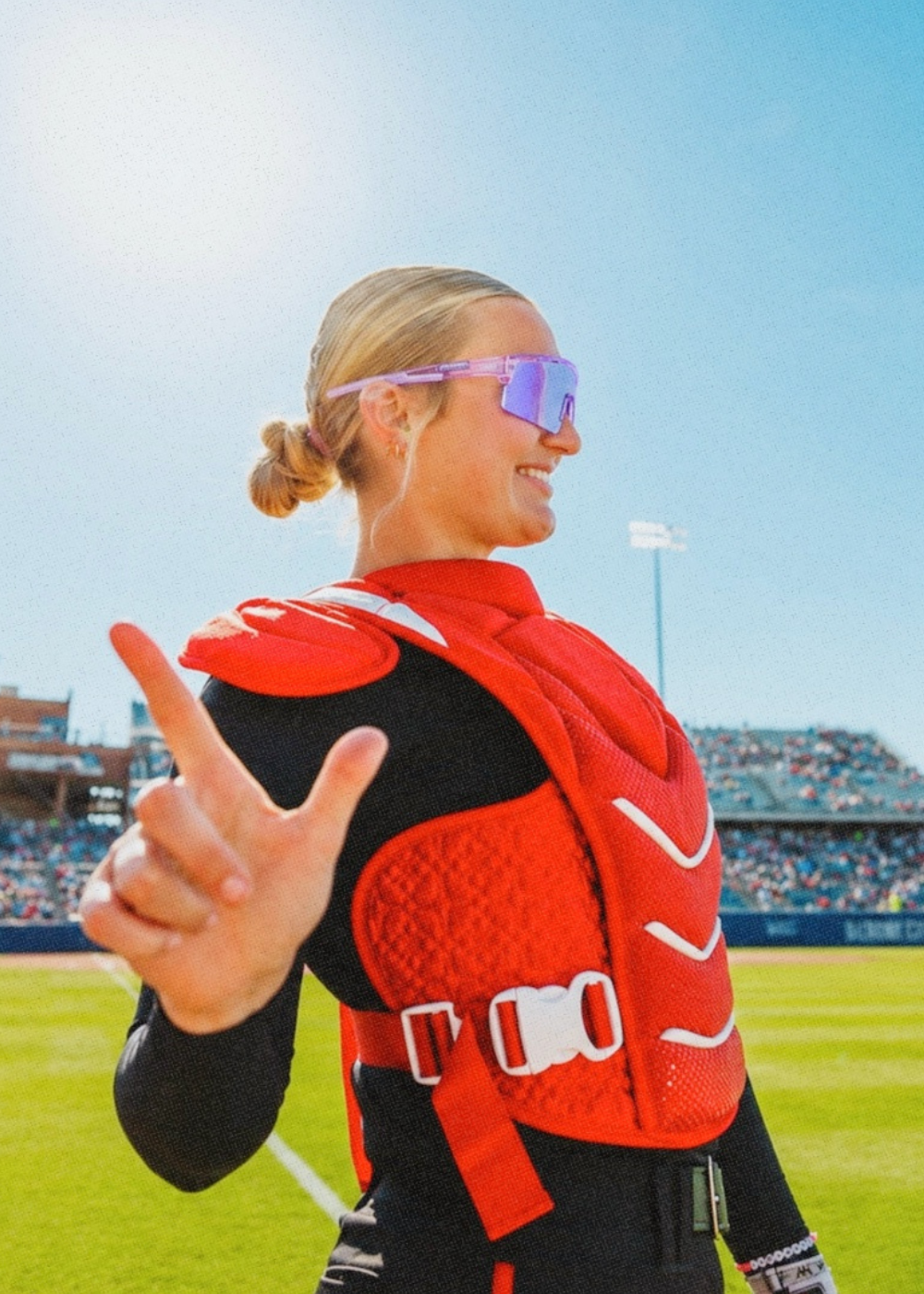 Person in a red catcher's vest on a baseball field giving a thumbs-up gesture wearing lavender purple solaro shades performance baseball sunglasses.