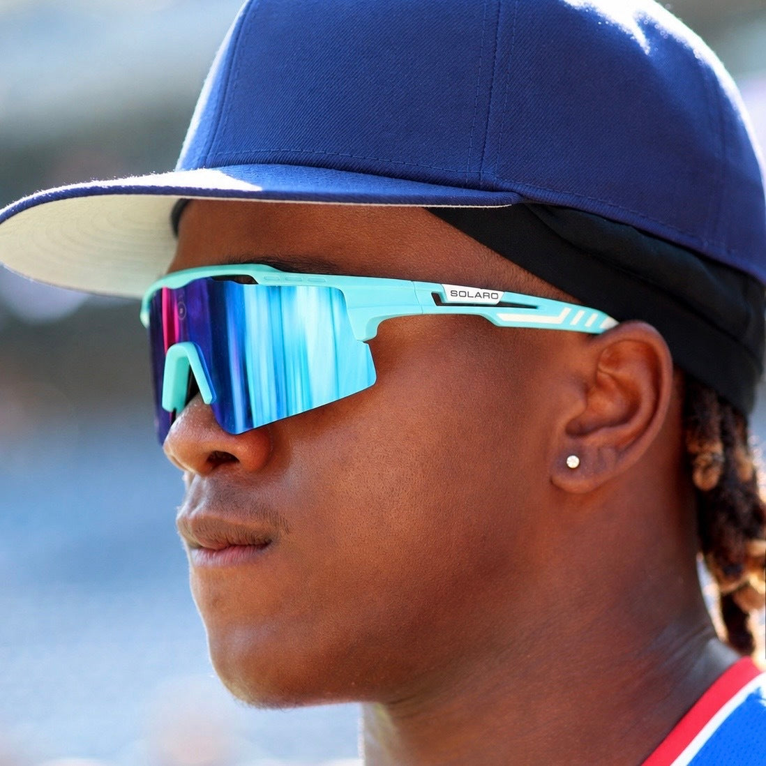 A close-up side profile of Jesus Made wearing reflective blue and purple mirrored sports solaro sunglasses sunglasses with a light blue frame, a blue baseball cap, and a black headband.