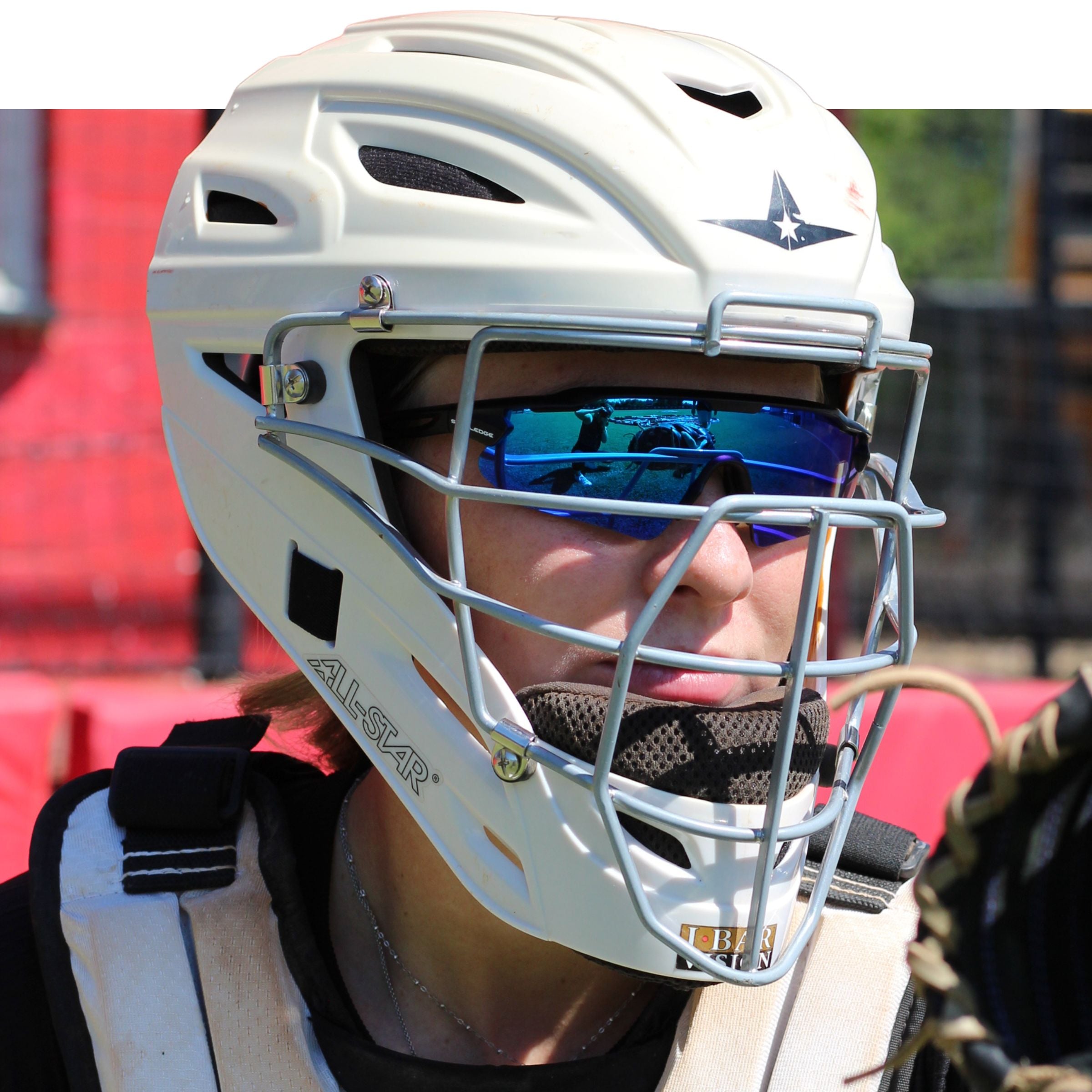 Person wearing a white catcher's helmet with a face mask and blue Solaro Shades baseball sunglasses, standing in front of red bleachers.