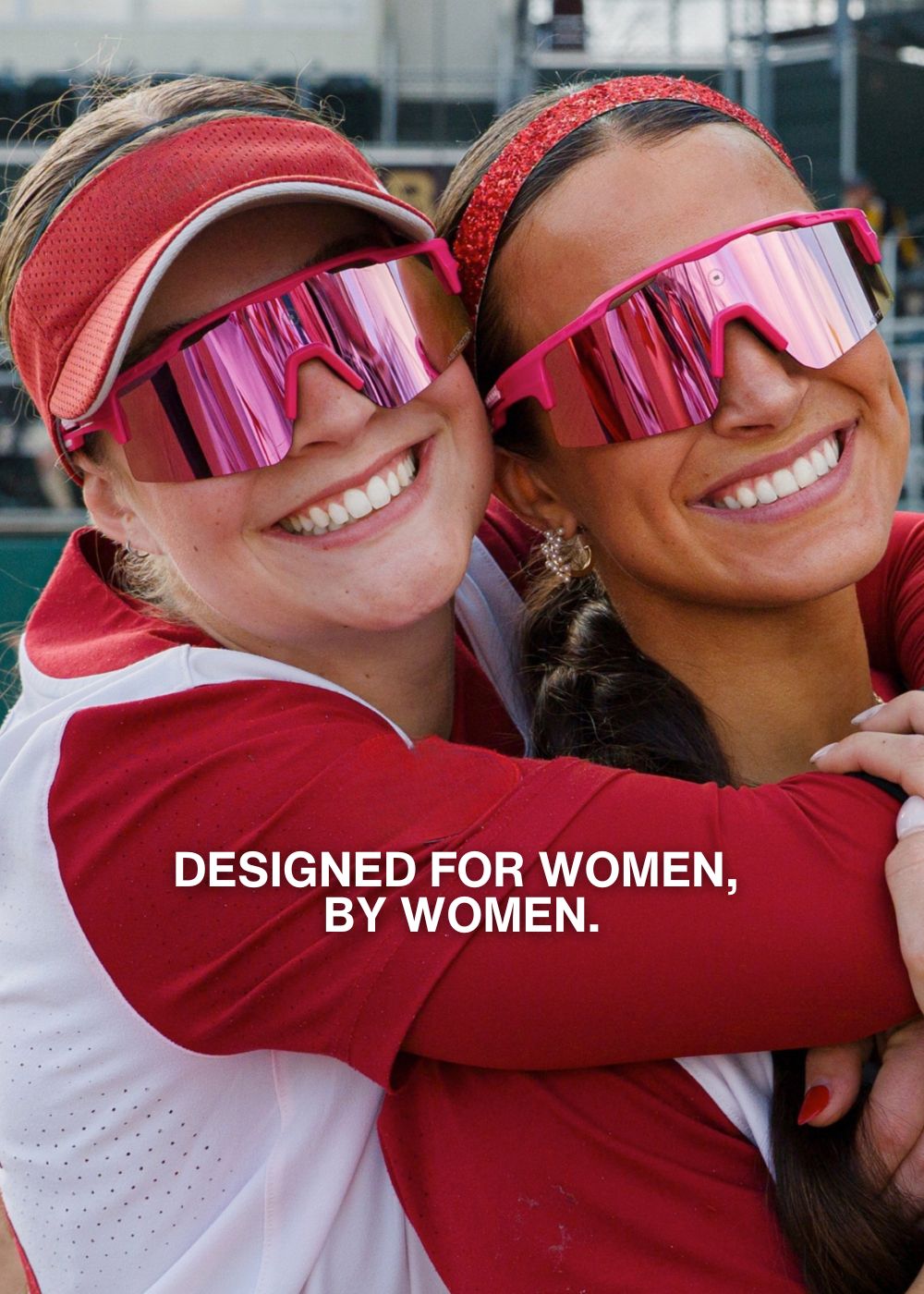 Two women wearing pink Solaro Shades softball sunglasses and red visors hugging, with text 'Designed for women, by women' on a blurred background.