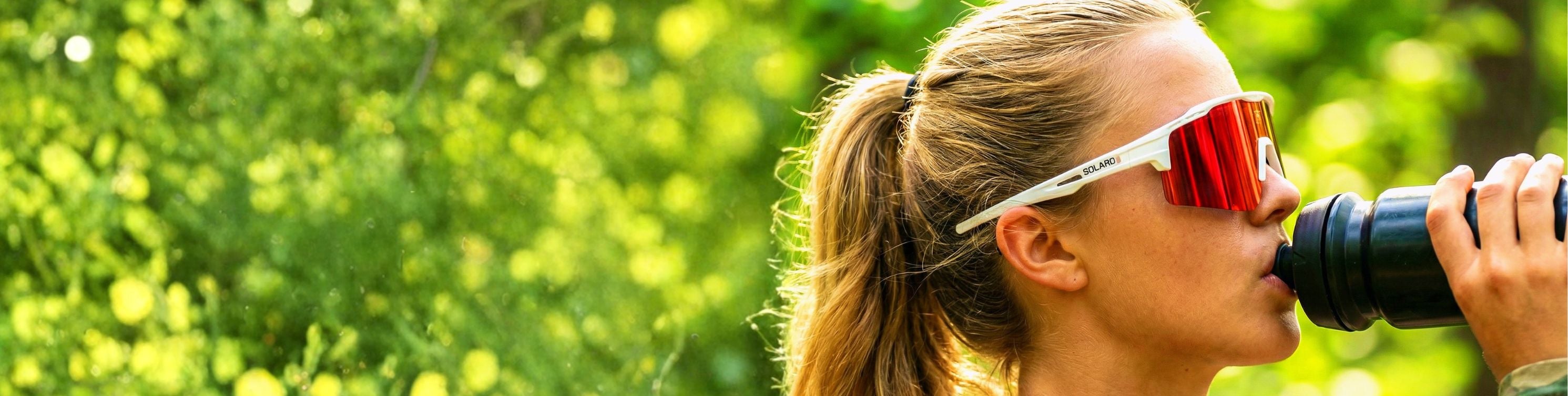 Person drinking from a water bottle with Solaro Shades biking sunglasses on, surrounded by greenery.