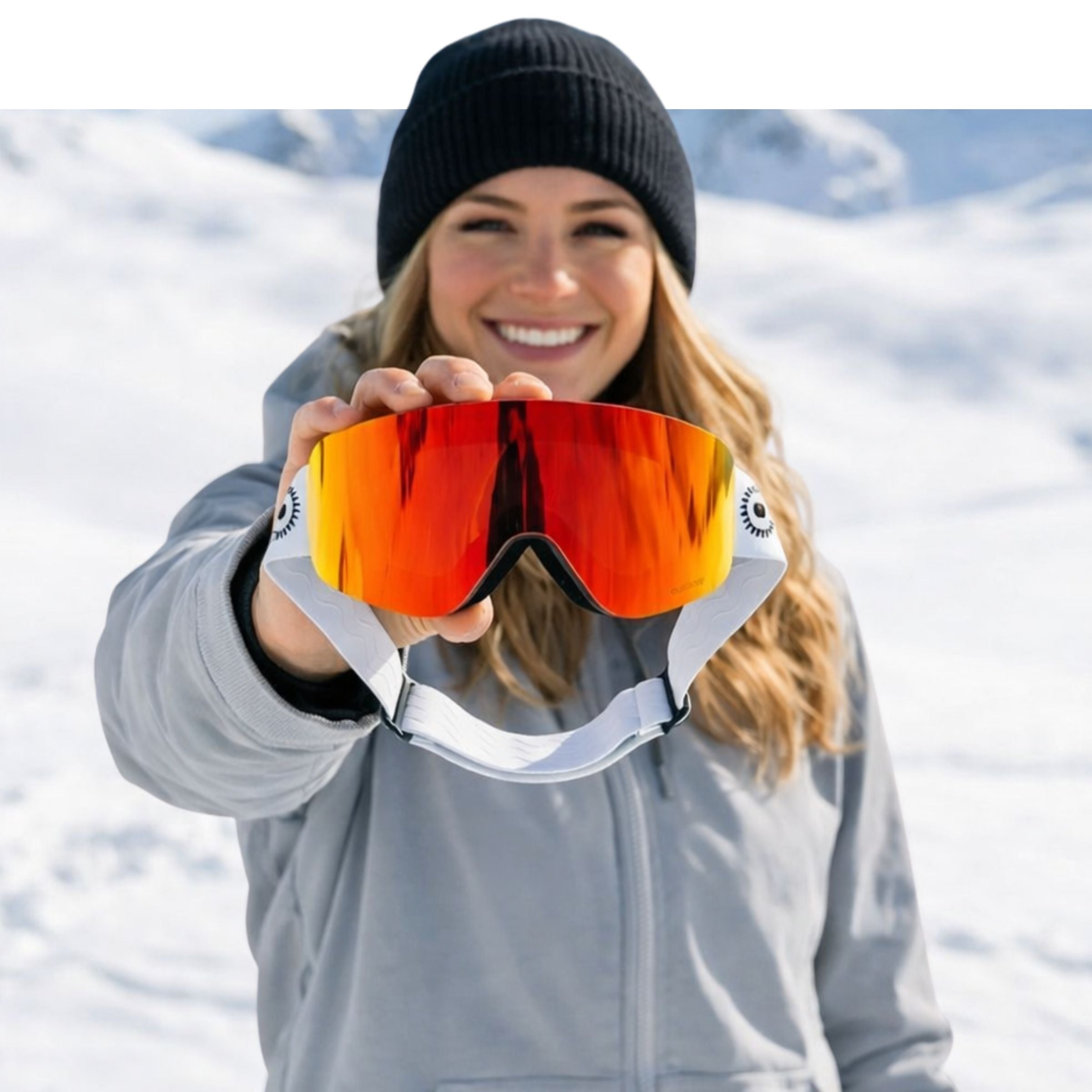 Woman holding up a pair of orange Solaro Shades ski goggles with a snowy mountain background