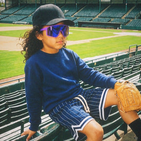 Child sitting on a stadium bench wearing a blue shirt, black cap, and Solaro jamberry jr. purple Pluto sunglasses with a baseball glove.