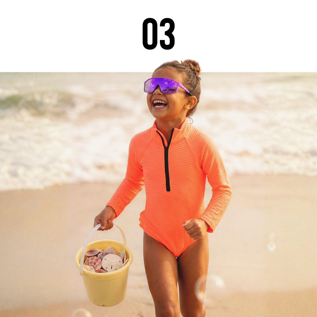 Child in Solaro Shades kids sunglasses and an orange swimsuit with sunglasses holding a bucket on a beach