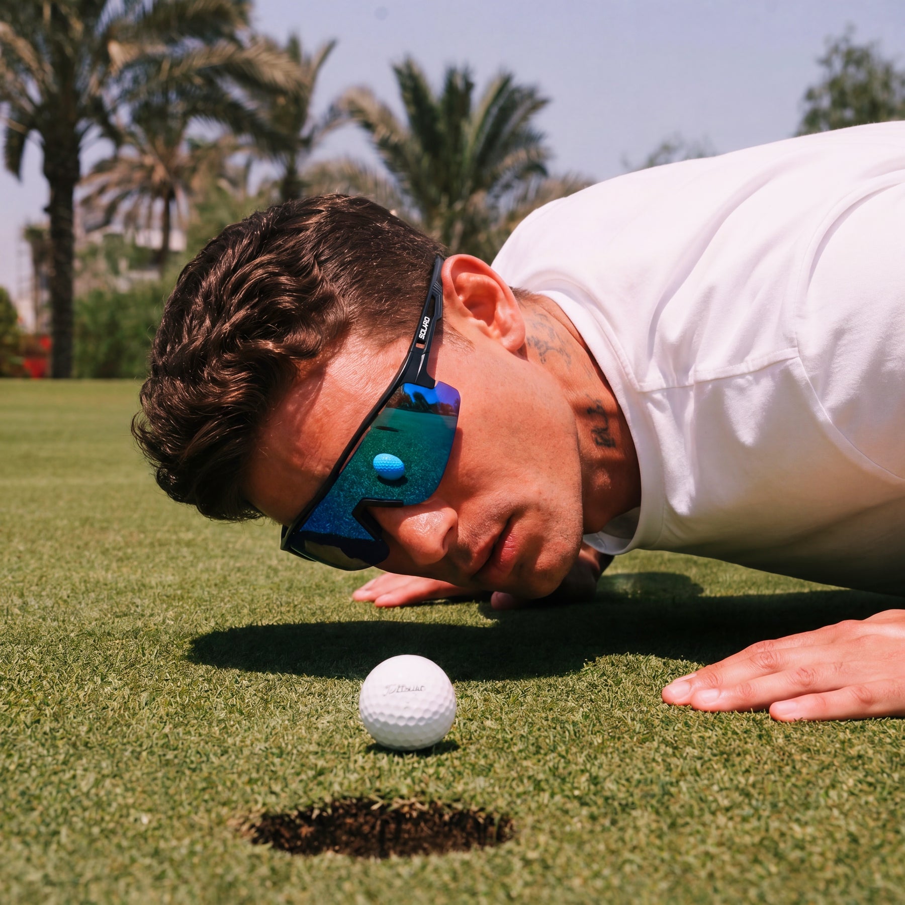 A man in a white shirt and blue mirrored solaro shades sunglasses leans down closely to a golf ball on a golf course green, with palm trees and the sky in the background.