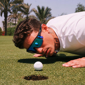 A man in a white shirt and blue mirrored solaro shades sunglasses leans down closely to a golf ball on a golf course green, with palm trees and the sky in the background.