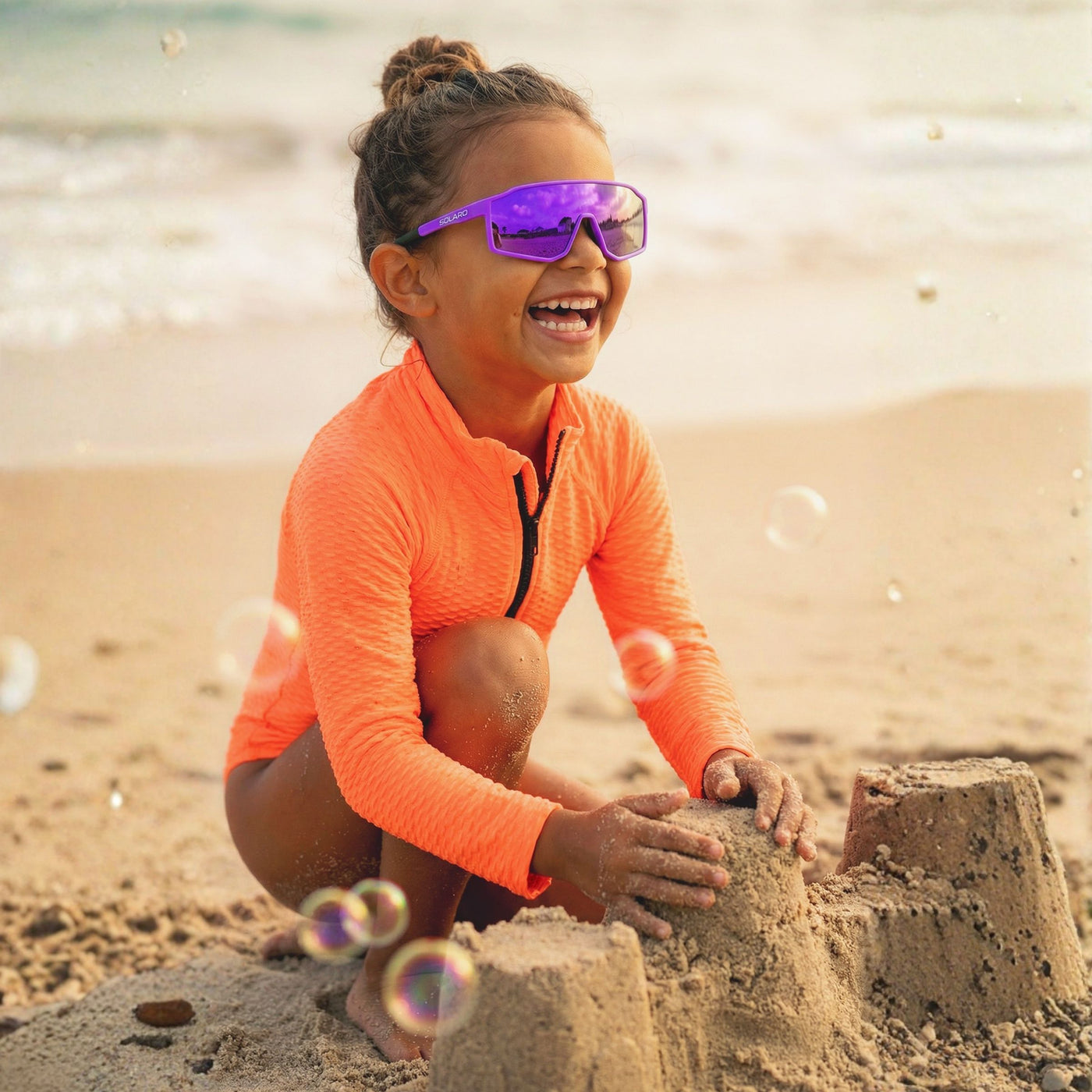 Child wearing orange shirt and Solaro Shades kids sunglasses that are purple sunglasses building a sandcastle on a beach.