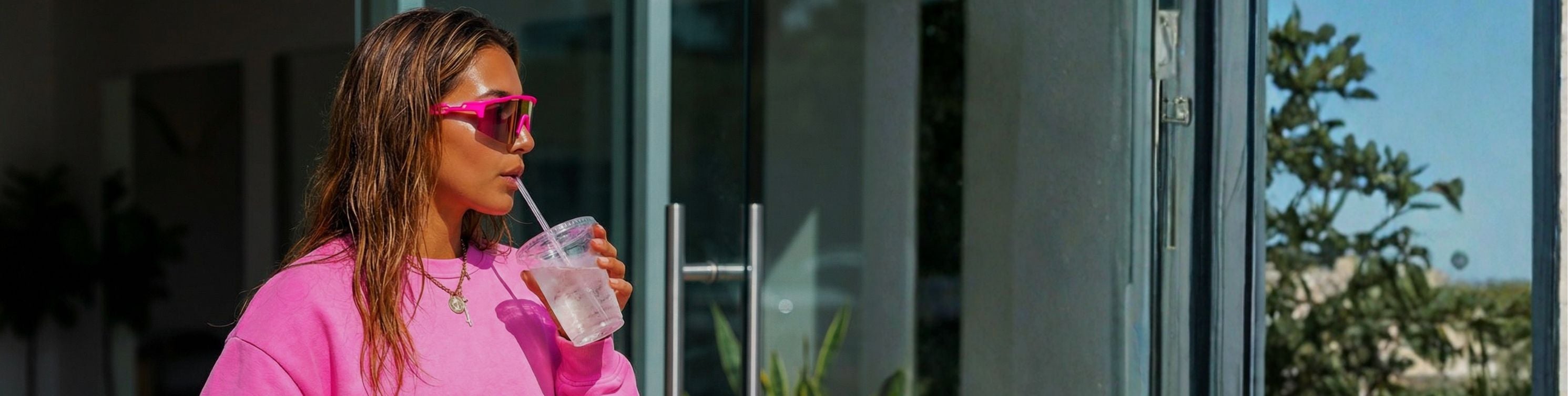 Woman in pink sweatshirt and sunglasses drinking from a straw outdoors wearing Solaro Shades Hot Pink sports sunglasses. 