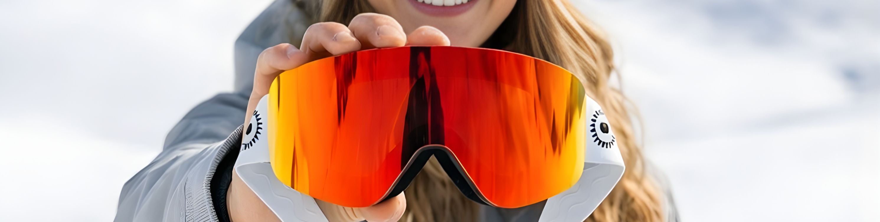 Woman holding up a pair of orange Solaro Shades ski goggles with a snowy mountain background