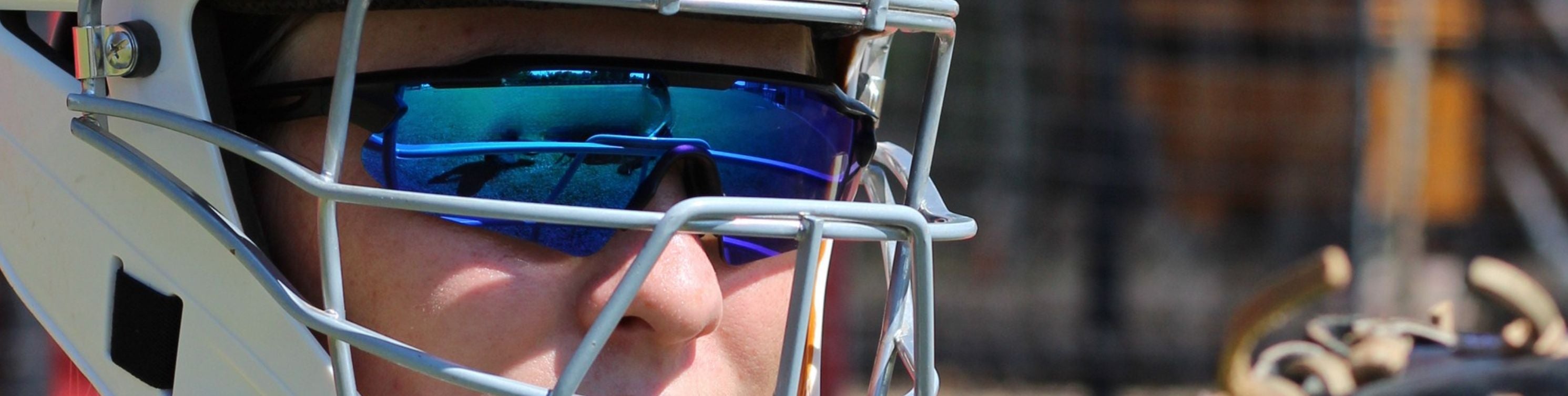 Person wearing a white catcher's helmet with a face mask and blue Solaro Shades baseball sunglasses, standing in front of red bleachers.