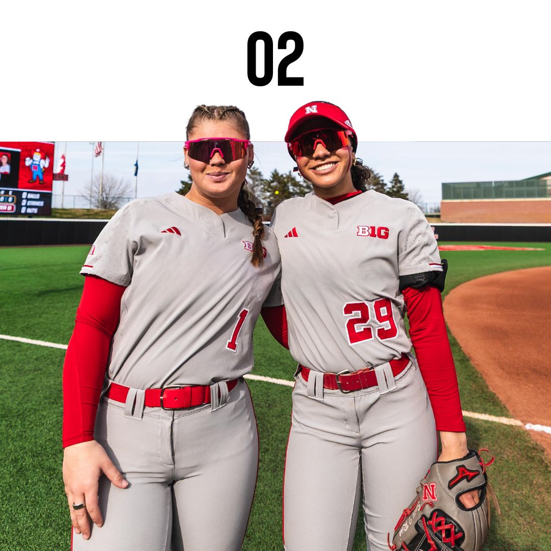 Sammie Bland of Nebraska softball wearing softball sunglasses and gray and red uniforms on a baseball field with a scoreboard in the background.